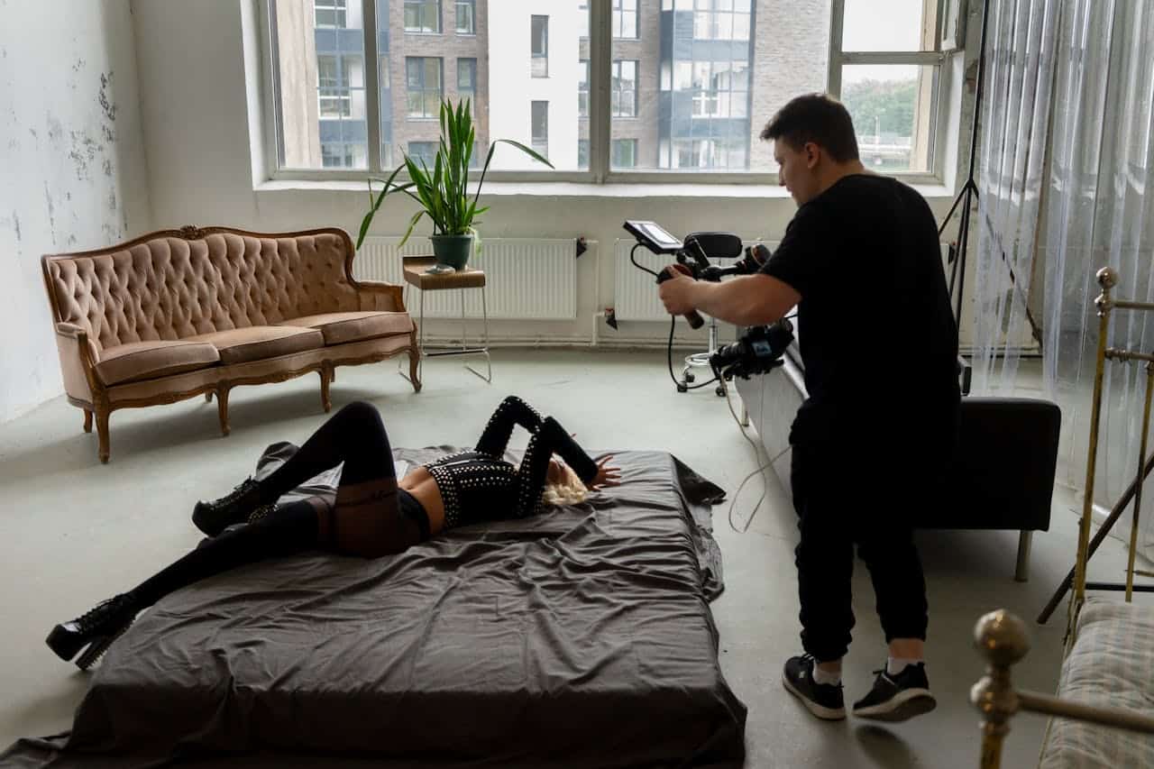 Fotograf richtet eine Kamera auf eine Frau in stilvollem Outfit, die auf einem Bett in einem hellen Studio mit vintage Möbeln posiert.