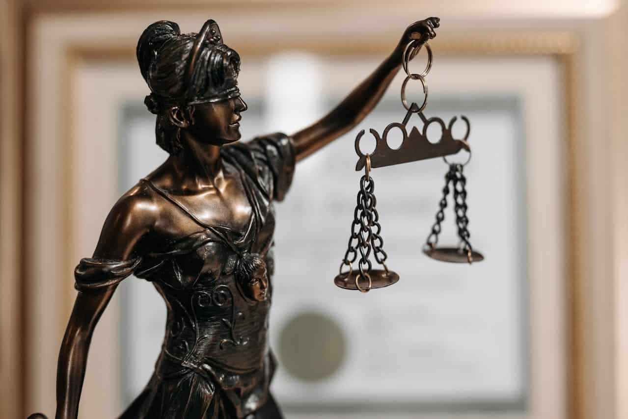 Nahaufnahme einer Justitia-Statue mit Waage und verbundenen Augen, vor einem unscharfen Zertifikathintergrund.